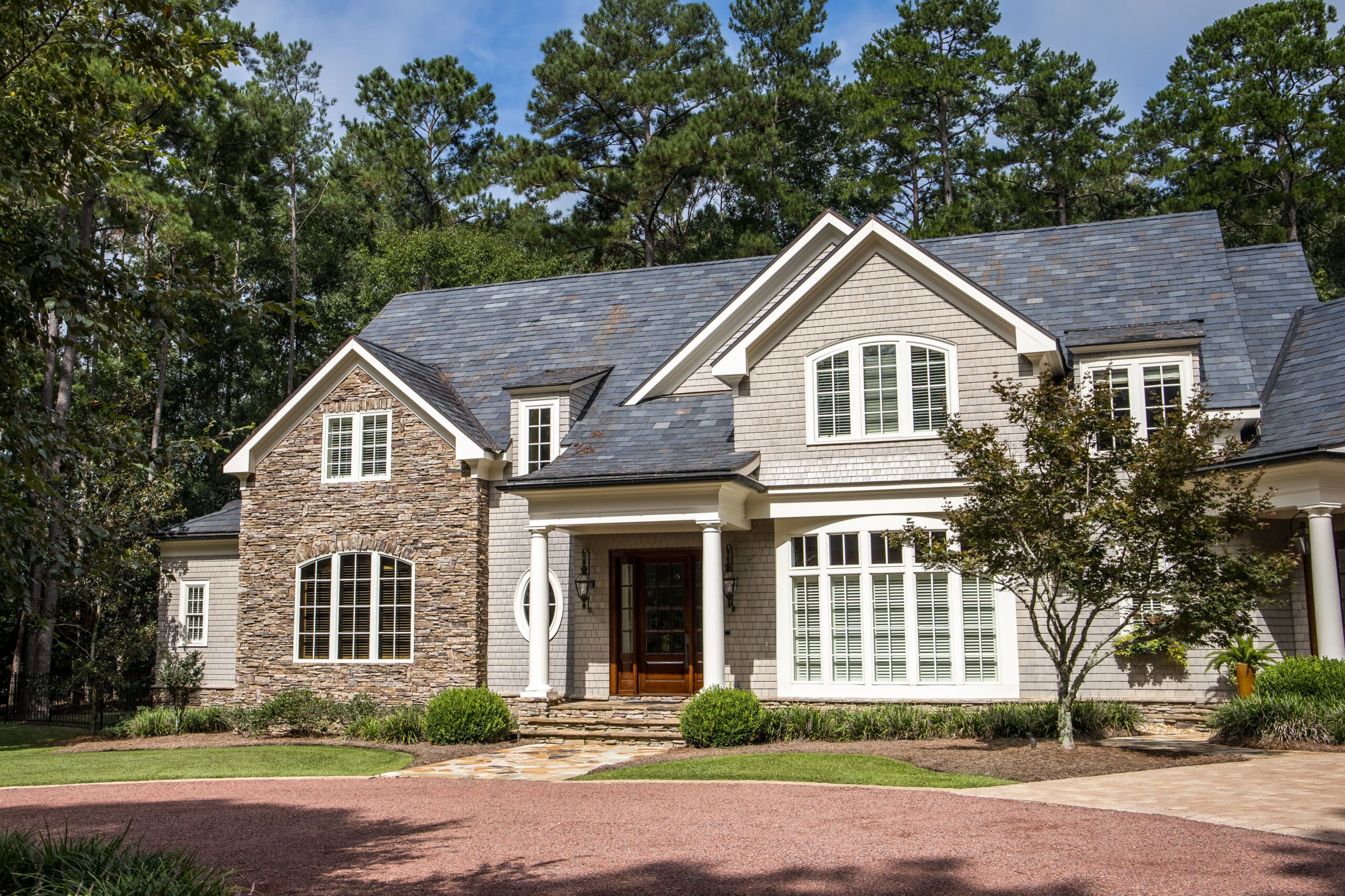 Front view of large estate home in the south with a gravel driveway and lots of windows. house made of brick, stone and clapboard in a cape cod style